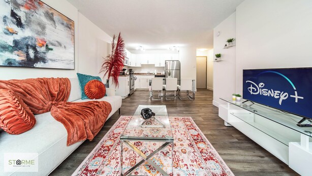 A modern living room with a sofa, a glass coffee table, a flat-screen TV, and a kitchen in the background.