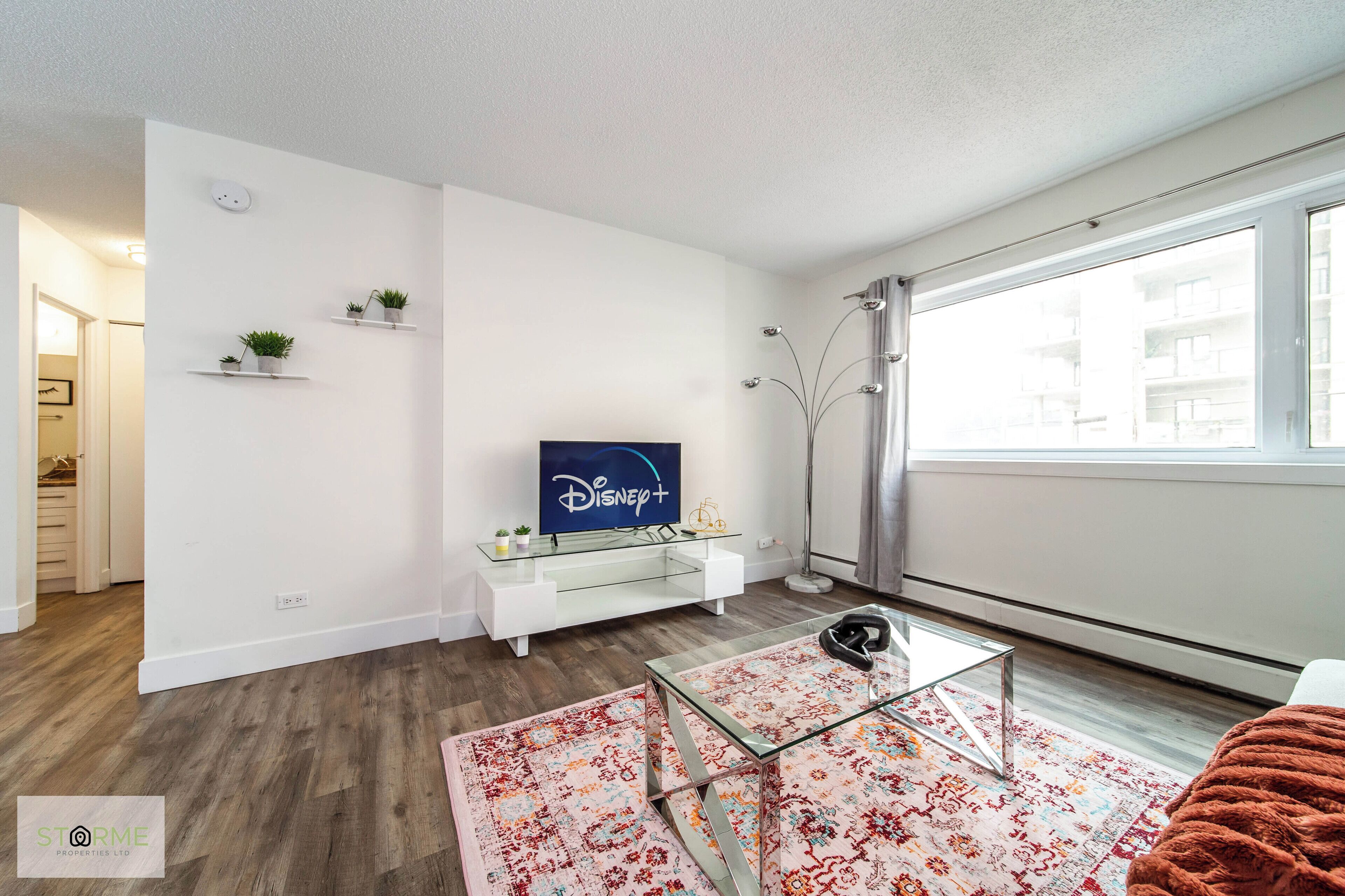 A modern living room with a flat-screen TV, white furniture, a glass coffee table, and a large window with curtains.