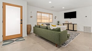 A modern living room with a green sofa, a large window with blinds, a flat-screen TV mounted on the wall, and a decorative basket.