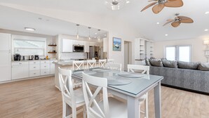 Et moderne køkken med hvide skabe, et spiseområde med et glasbord og hvide stole, samt en grå sofa i opholdsstuen.
