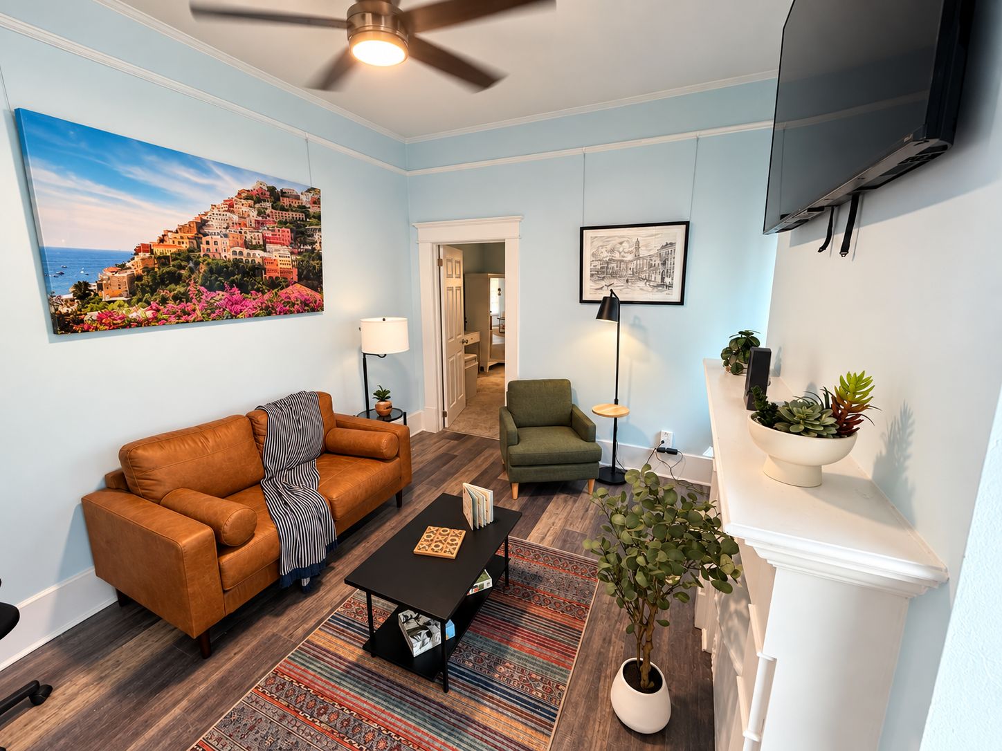 A living room with a sofa, armchair, coffee table, and a painting of a coastal town.