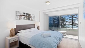 A modern bedroom with a large bed, a bedside table with a lamp, and a view of a snowy landscape through a large window. - Goodwin Towers 10, Burleigh Heads. (Gold Coast)