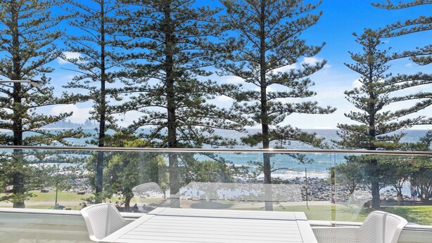 A balcony with a table and chairs, overlooking a beach with trees and a distant city skyline. - Goodwin Towers 10, Burleigh Heads. (Gold Coast)