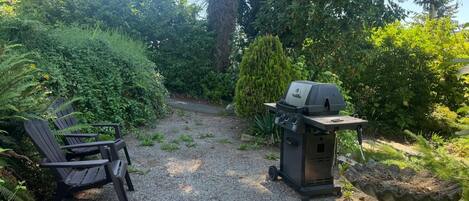 A gravel path leading to a barbecue grill and Adirondack chairs in a garden setting.
