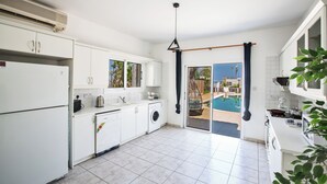 A modern kitchen with white cabinets, a built-in washing machine, and a view of a swimming pool. - Protaras Fig Tree Bay Villa Jv4 (Paralimni)