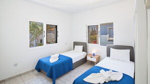 A hotel room with two single beds, each with a blue bedspread and white pillows, a small bedside table with a lamp, and a window with a view of palm trees and a building. - Protaras Fig Tree Bay Villa Jv4 (Paralimni)