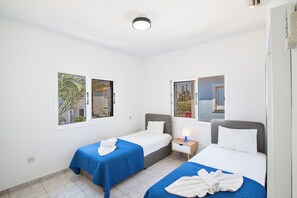 Habitación de hotel con dos camas individuales, cada una con cubrecama azul y almohadas blancas, una mesita de noche con lámpara, y una ventana con vistas a palmeras y un edificio.