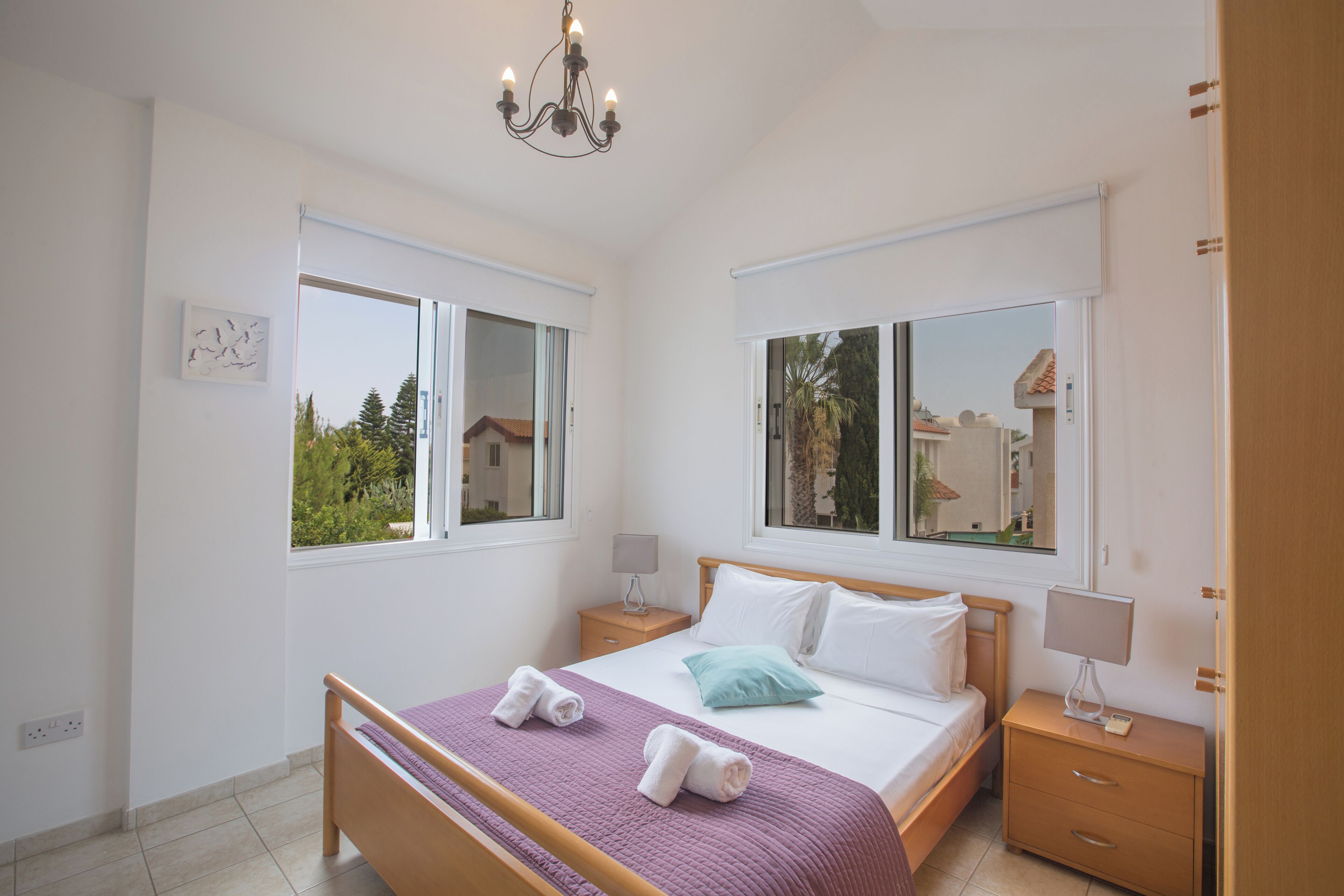 A bedroom with a bed, two bedside tables, a chandelier, and large windows.