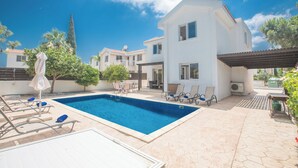 A swimming pool with lounge chairs and umbrellas, surrounded by a white building with a tiled roof. - Protaras Holiday Villa Ma14 (Paralimni)