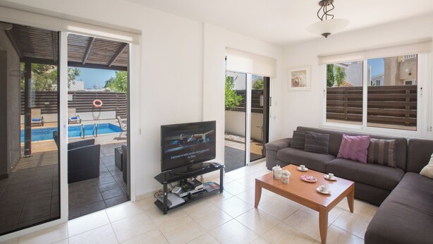 A living room with a television, a coffee table, and a sofa. There is a pool visible through the glass doors. - Protaras Holiday Villa Ma14 (Paralimni)
