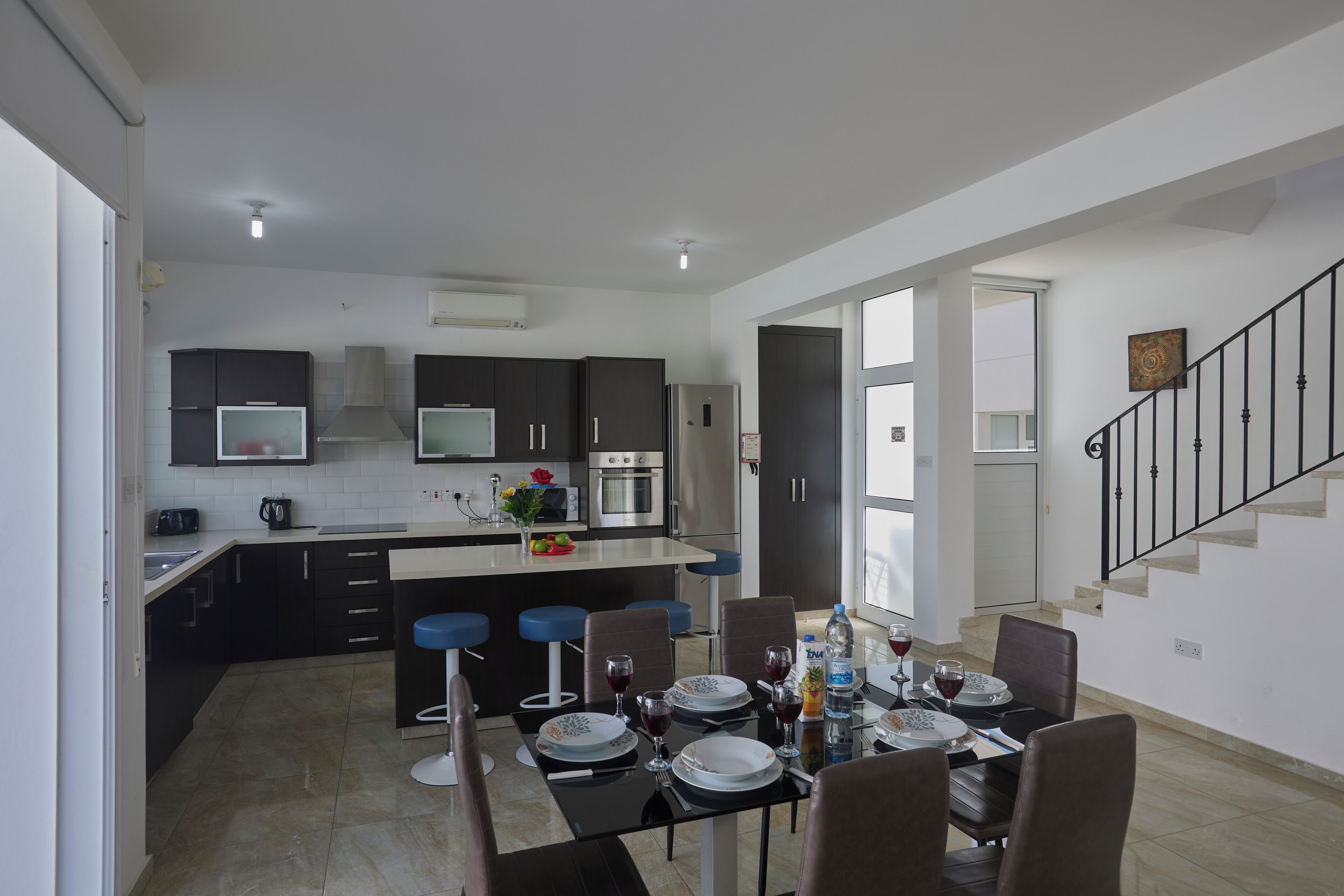 A modern kitchen with dark cabinets, a central island, and bar stools. There is a dining area with a table set for four, and a staircase with a black railing.