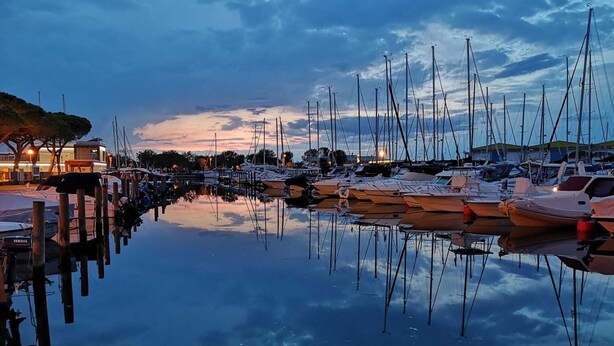 A marina with multiple boats docked, calm water reflecting the boats and sky, and buildings in the background. - Villa With Pool, Garden and Private Parking by Beahost Rentals (Caorle)
