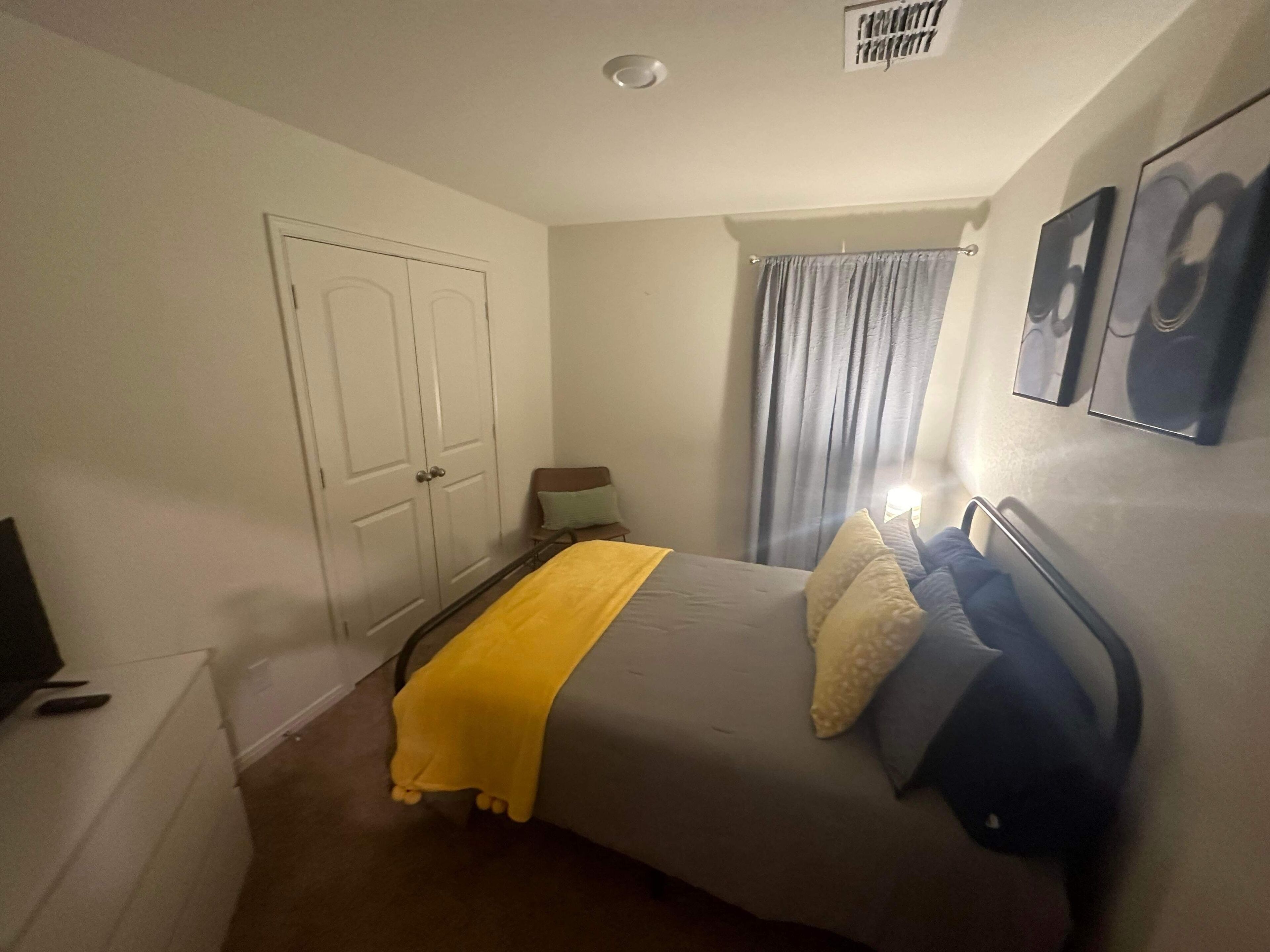 Quarto com cama, mesa de cabeceira, cadeira, guarda-roupa e janela com cortinas.