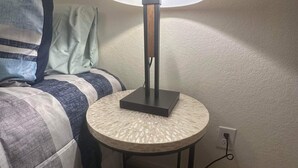 Un lit avec une couette à carreaux et des oreillers, une petite table ronde avec une lampe, et une prise électrique sur le mur.