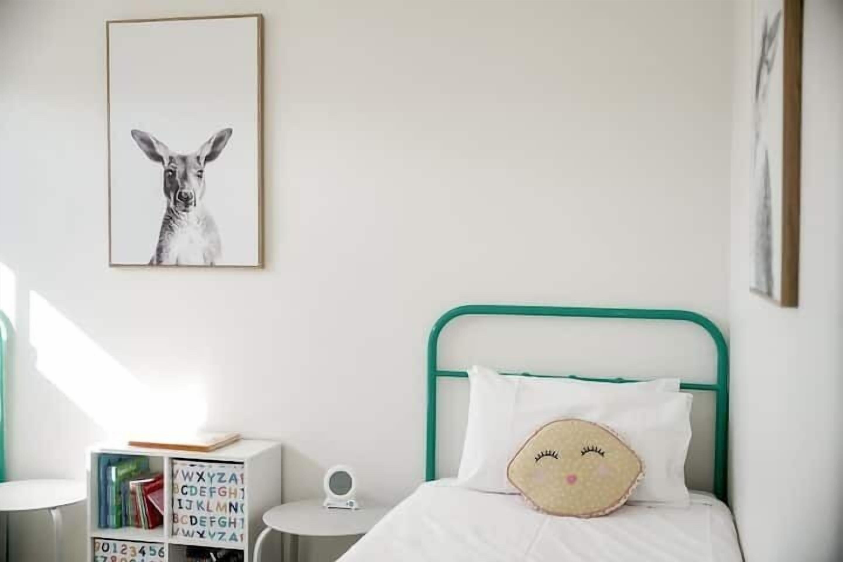 Un dormitorio con estructura de cama metálica verde, ropa de cama blanca, una estantería con libros, un cuadro en la pared y una mesita de noche con lámpara.