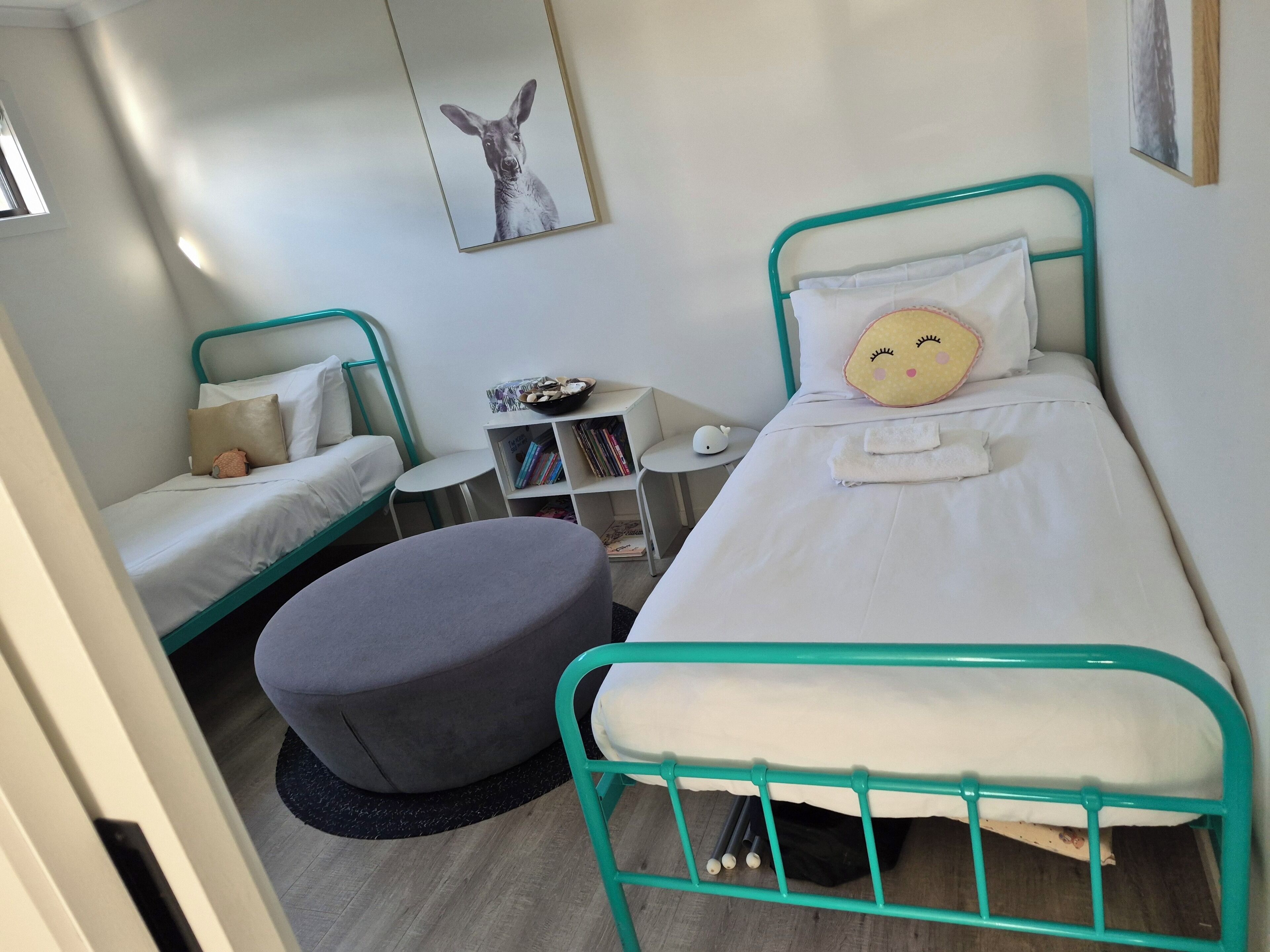 A small, clean room with two single beds, a gray ottoman, a small table with a bowl of snacks, and a bookshelf.
