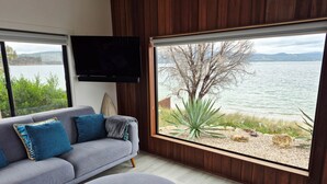 A modern living room with a grey sofa, blue cushions, and a round table with a book. A large window offers a view of a lake and trees.