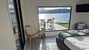 A modern bedroom with a large window overlooking a lake and mountains. It includes a bed, a chair, and a view of the outdoors.