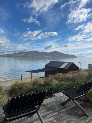 A wooden deck with two lounge chairs overlooking a lake, a small building with a sloped roof, and a distant mountain range under a partly cloudy sky.