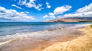 A beach with clear blue water, sandy shore, and distant mountains under a partly cloudy sky.