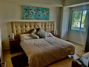 Un dormitorio con una cama grande, cabecera, dos mesitas de noche, un cuadro y una ventana con vistas a zonas verdes.