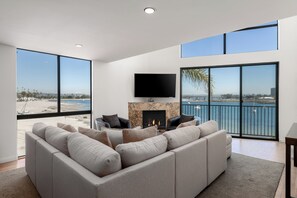 Un salon moderne doté d’un grand canapé d’angle gris, d’un téléviseur à écran plat fixé au-dessus d’une cheminée et d’un balcon offrant une vue sur la mer.