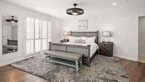 A bedroom with a wooden bed, two bedside tables with lamps, a bench, a large window with blinds, and a ceiling light fixture. - Large Waterfront | Private Roof Deck | Fireworks View (San Diego)