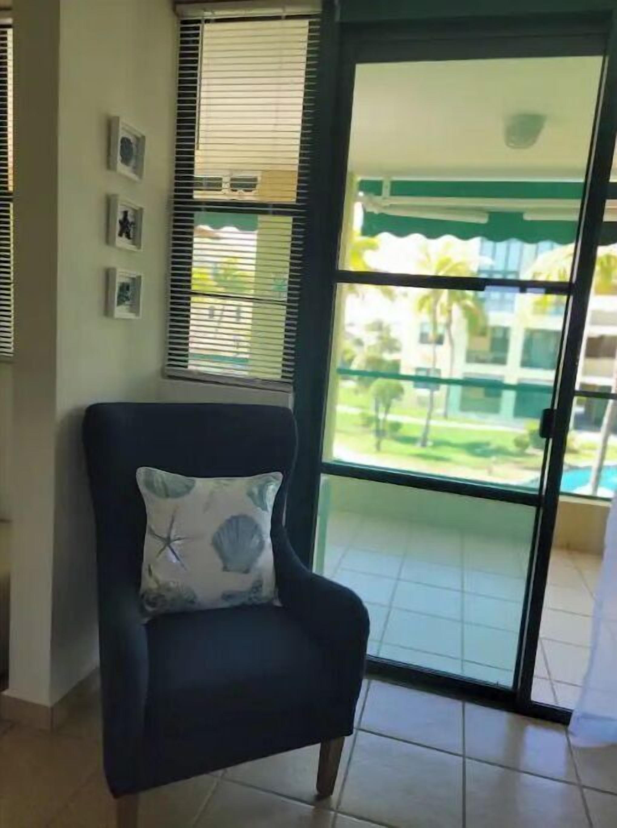 Een kamer met een donkergroene fauteuil met een decoratief kussen, een raam met jaloezieën, en uitzicht op een zwembad en groen.