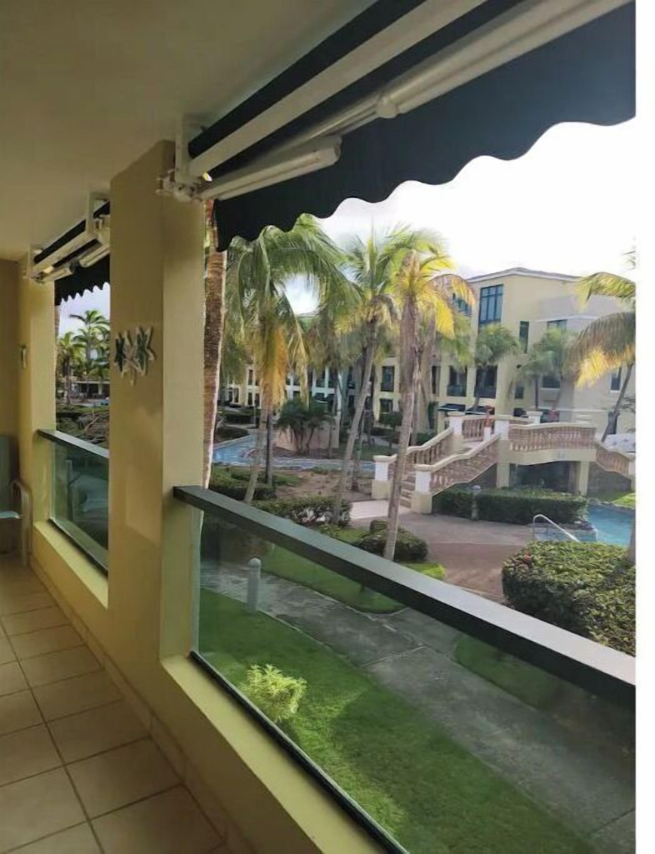 A balcony with a view of a pool area, palm trees, and a building with a staircase.