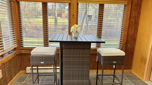 Ein Eckzimmer mit dunklem Korbstuhl-Tisch und zwei passende Stühle, einer kleinen Vase mit Blumen sowie großen Fenstern mit Jalousien.
