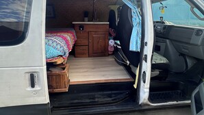 Ein Van mit offener Rückwand, der ein Bett, eine kleine Holzschublade und eine Küchenzeile zeigt.