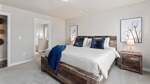 Un dormitorio con estructura de cama de madera, ropa de cama blanca y una manta azul. Hay una mesita de noche con una lámpara y un cuadro en la pared.