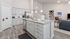 Una cocina moderna con armarios blancos, encimera de granito y electrodomésticos de acero inoxidable. Hay un rincón de comedor con un sofá gris y un televisor de pantalla plana.