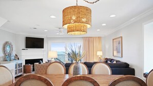 A dining area with a wooden table and chairs, a large woven pendant light, a flat-screen TV above the fireplace, and a view of the outdoors through large windows. - Gulf Front-Unobstructed Views! Book Now! (Port St. Joe)