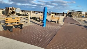 A wooden boardwalk with benches and a blue structure, leading to a sandy beach area with wooden fences and a clear sky.