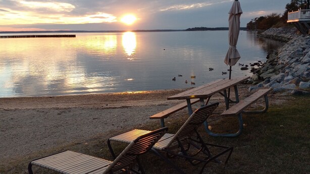 Zona de picnic junto al lago con bancos y una sombrilla, con vistas a un cuerpo de agua tranquilo y un atardecer.
