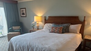 Una cama bien hecha con edredón blanco y almohadas, cabecera de madera, lámpara de noche, mesita de noche con lámpara y planta en maceta, y una silla con cojín.