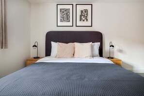 A neatly made bed with a dark headboard, two bedside tables with lamps, and two framed abstract artworks on the wall.