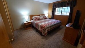 Un dormitorio con cama, mesitas de noche, lámpara, ventana con cortinas y un mueble para televisor.