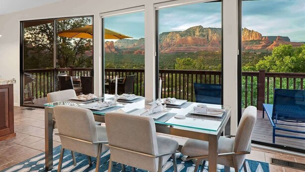 A dining area with a glass table and chairs, large windows with a view of red rock formations, and a wooden deck outside. - Best Panoramic Views in Sedona, Hot Tub + Firepit! (Sedona)