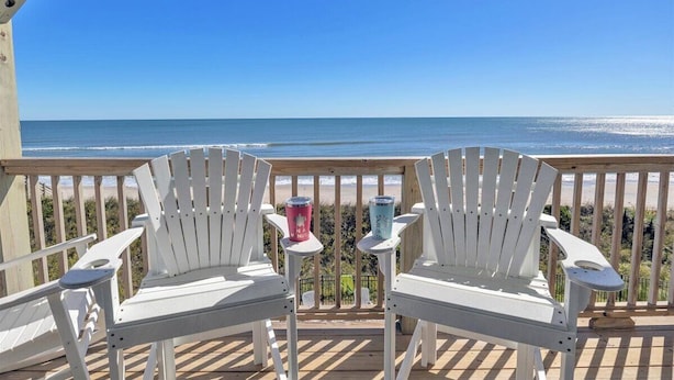 Eine Holzterrasse mit weißen Adirondack-Stühlen, die auf einen Strand ausgerichtet sind.