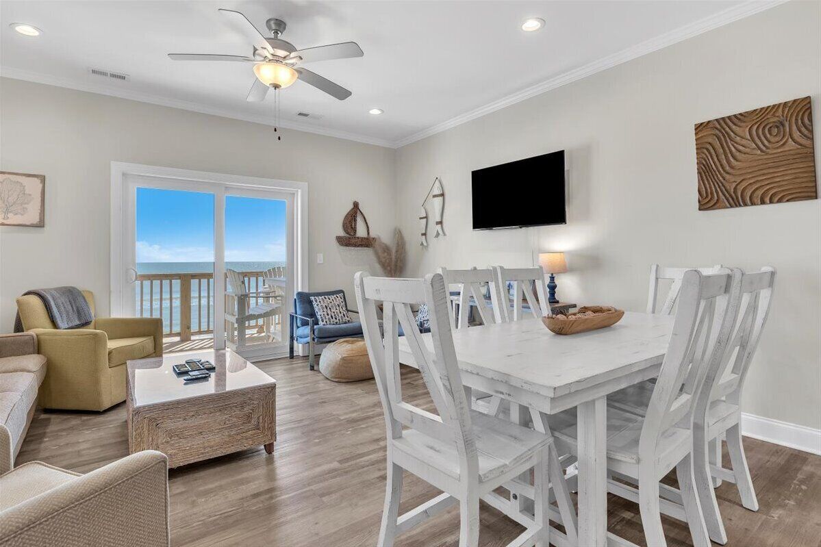 A spacious living room with a dining area, large windows, a ceiling fan, and a flat-screen TV mounted on the wall.