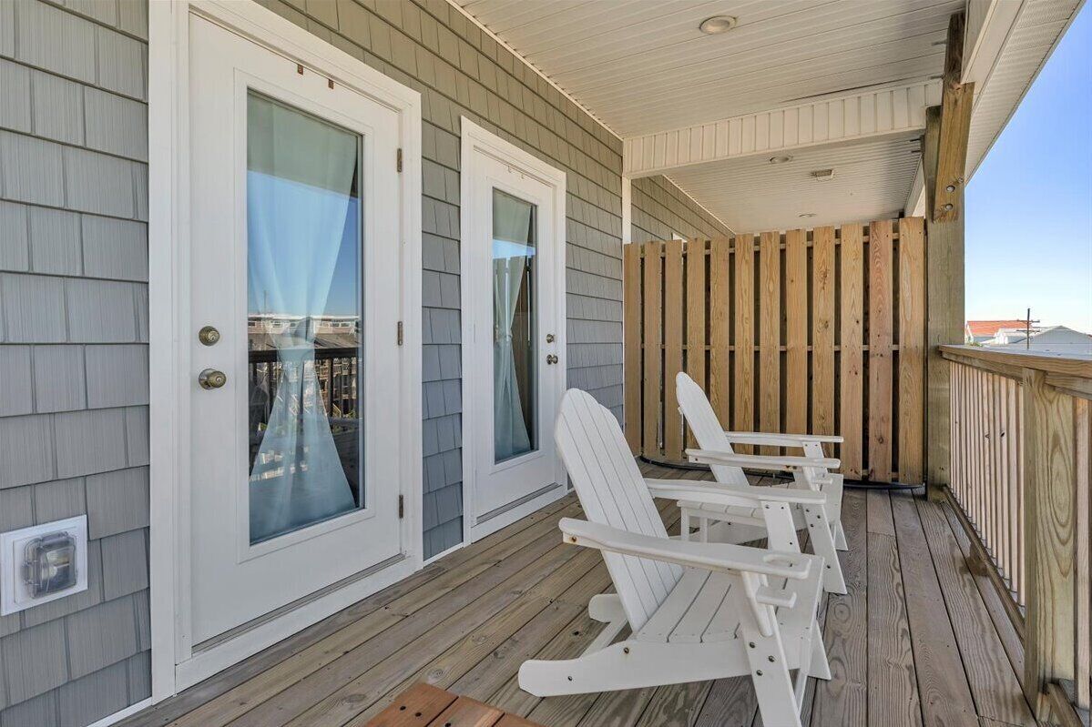 A covered porch with two sliding glass doors, wooden deck, and white Adirondack chairs.