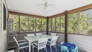 A covered patio with a glass table and chairs, a ceiling fan, and a view of trees through large windows. - Blue Skies - Bright, Spaciuos 4 BR oceanside home w/Private Pool and Hot Tub (Southern Shores)
