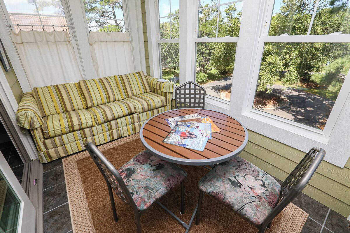 A sunroom with a striped sofa, a round wooden table with chairs, and large windows offering a view of greenery outside.