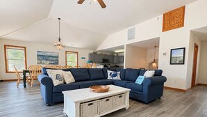 A spacious living room with a large sectional sofa, a white coffee table, and a dining area with wooden chairs. - Vitamin Sea - Recently updated 4 BR oceanside home w/Private Pool and Hot Tub (Duck)