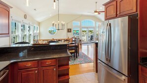 A spacious kitchen with wooden cabinets, a stainless steel refrigerator, and a dining area with a view. - 5202 Sunflower Siesta * 3 Min Walk to Beach (Kill Devil Hills)