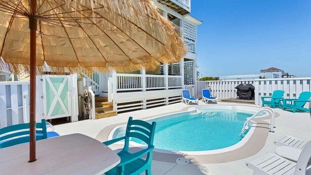 A pool area with a table and chairs, a thatched umbrella, and a view of a building in the background. - 5202 Sunflower Siesta * 3 Min Walk to Beach (Kill Devil Hills)