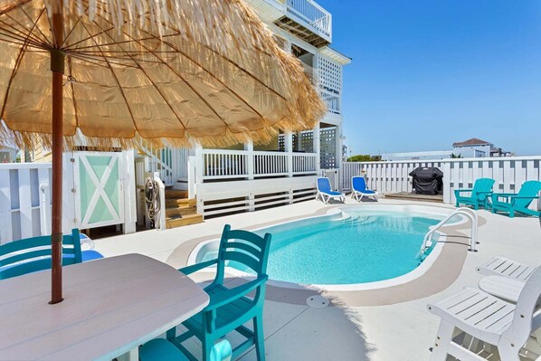 A pool area with a table and chairs, a thatched umbrella, and a view of a building in the background.