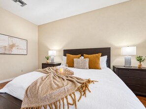 A hotel room with a large bed, two bedside tables, a nightstand, a wall-mounted air vent, and a framed picture on the wall.